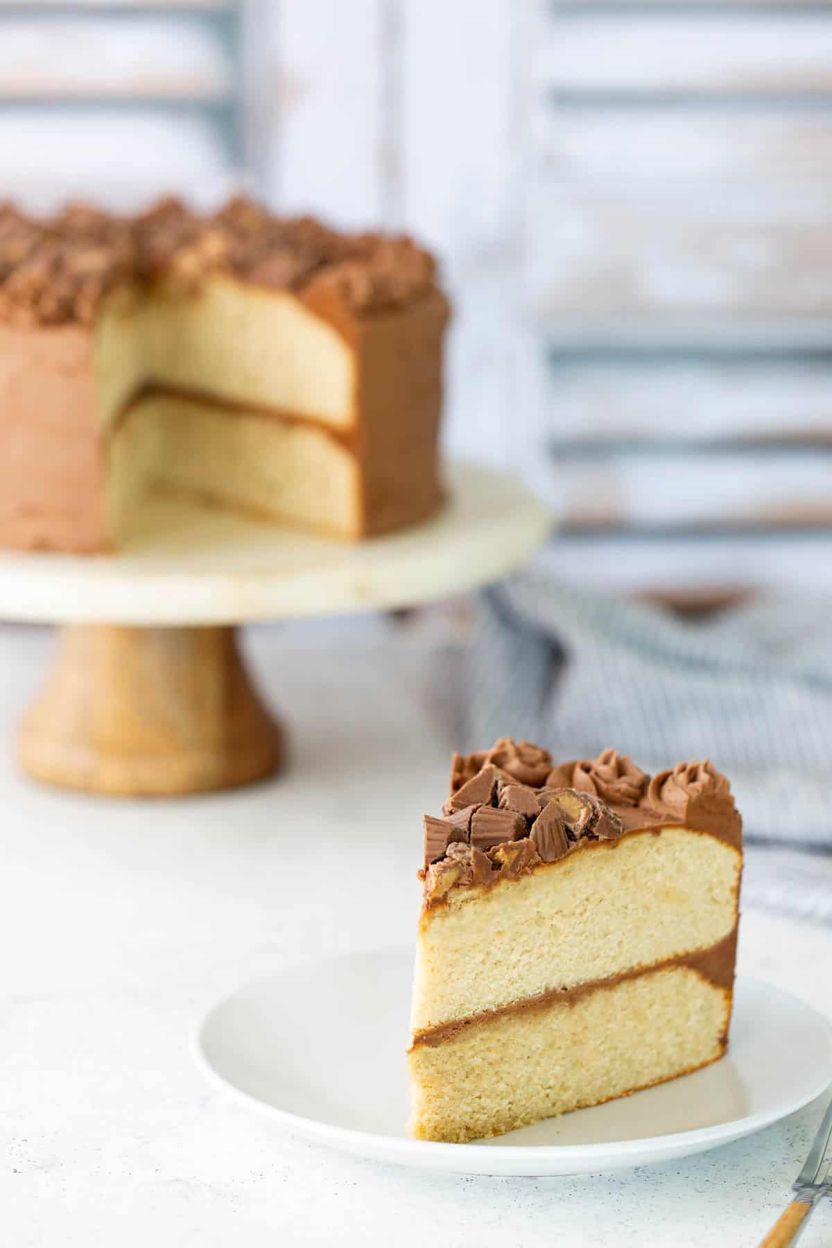 reeseheart A slice of two-layer vanilla cake with chocolate frosting sits on a white plate; the rest of the cake is on a cake stand in the background.
