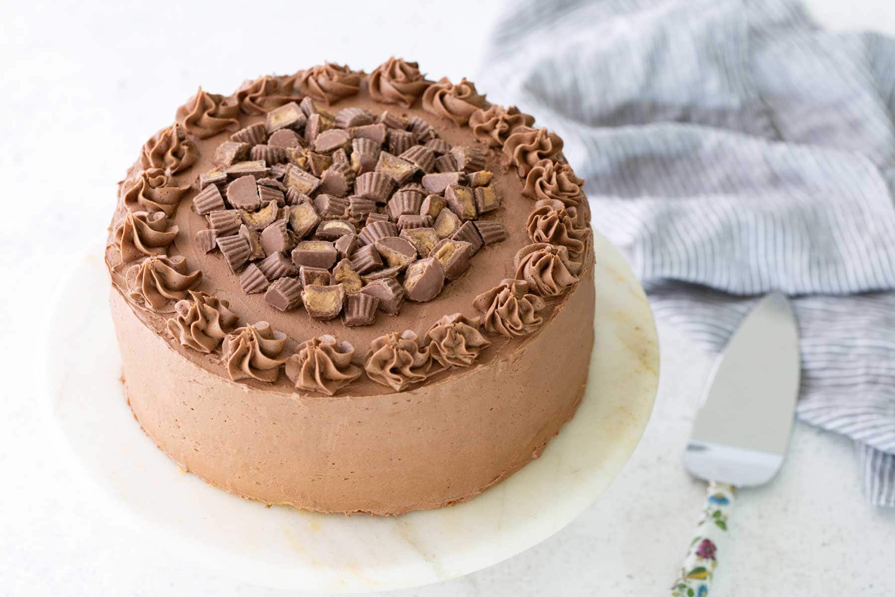 Chocolate cake with chocolate frosting, decorated with swirls around the edge and chopped peanut butter cups in the center, placed on a marble cake stand.