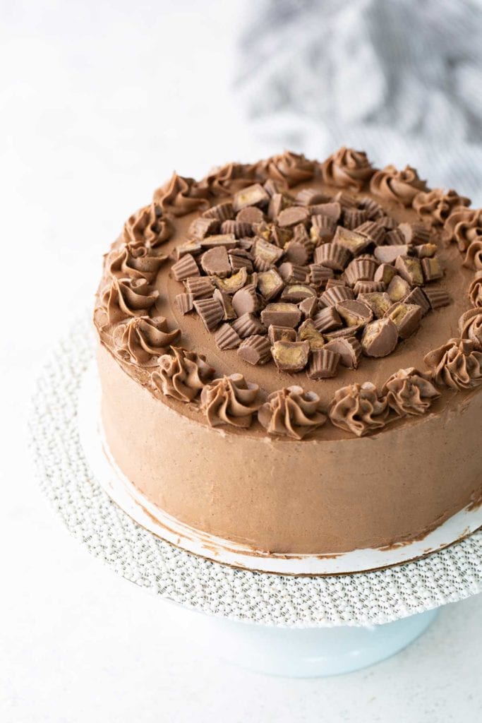 A round chocolate cake topped with chocolate frosting swirls and chopped peanut butter cups, displayed on a white cake stand.