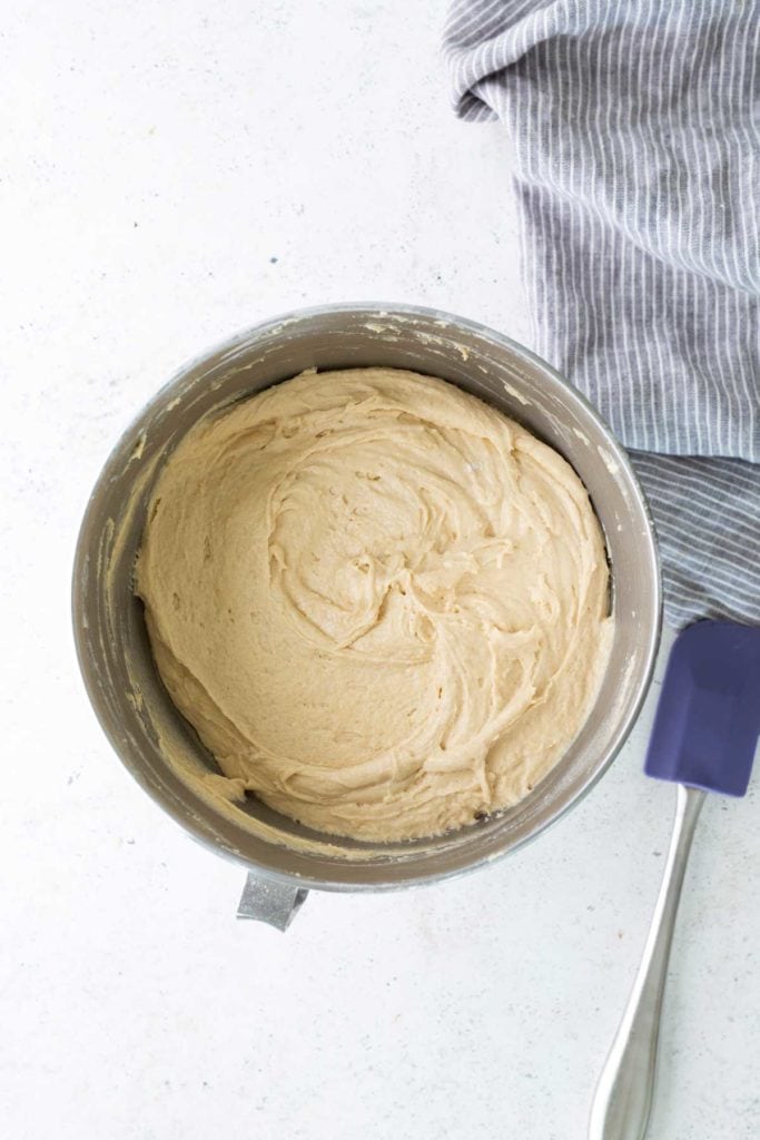 A metal mixing bowl filled with creamy, swirled batter sits beside a blue spatula and a gray striped towel on a light surface.