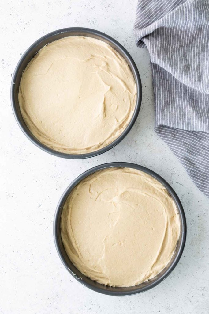Two round cake pans filled with smooth, unbaked cake batter sit on a white surface next to a gray and white striped cloth.