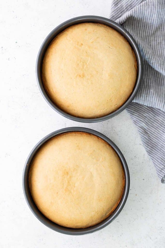 Two round, golden-brown cakes in metal pans on a white surface with a gray and white striped cloth partially visible.
