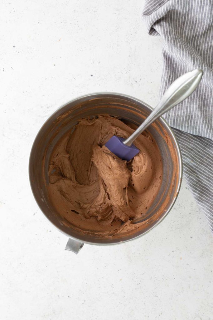 A metal mixing bowl filled with chocolate frosting and a purple spatula, placed on a white surface next to a gray striped cloth.