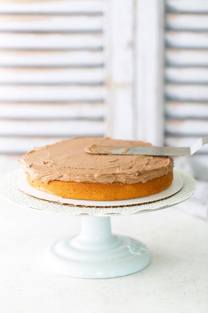 A single-layer yellow cake on a white cake stand being frosted with chocolate icing using an offset spatula.