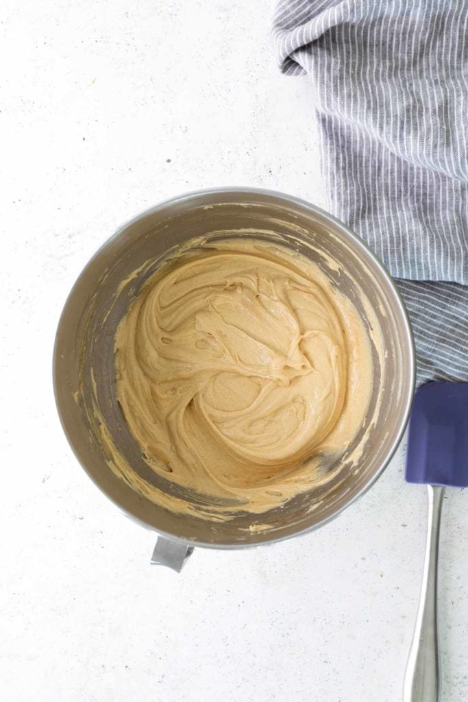 A metal mixing bowl containing light brown batter sits on a white surface next to a gray and white striped cloth and a purple spatula.