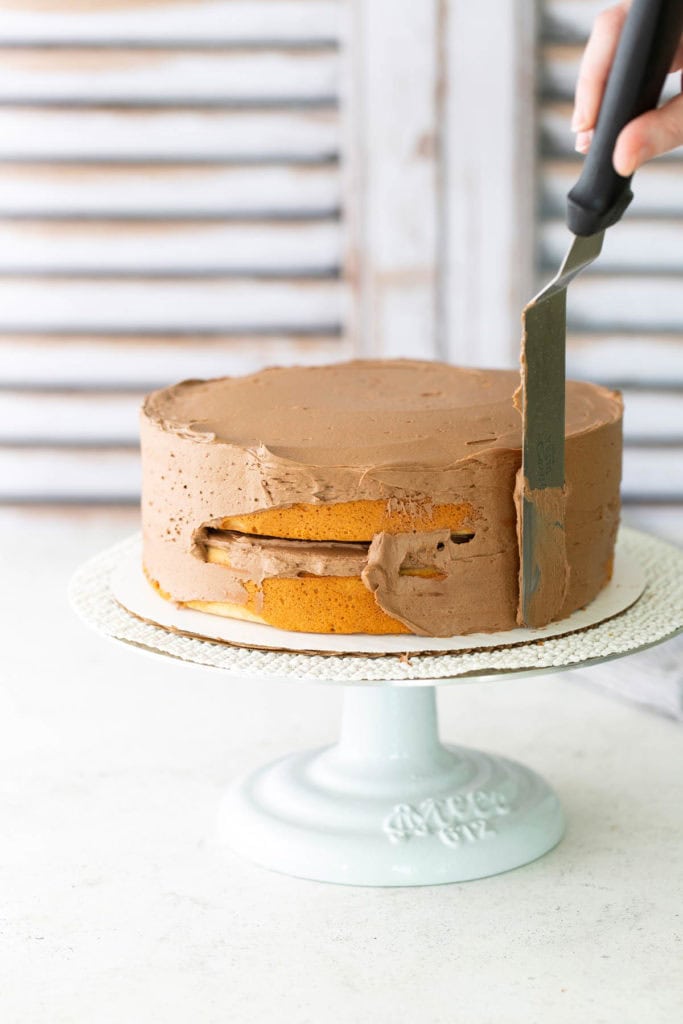 A hand uses an offset spatula to spread chocolate frosting on a round, yellow cake placed on a white cake stand.