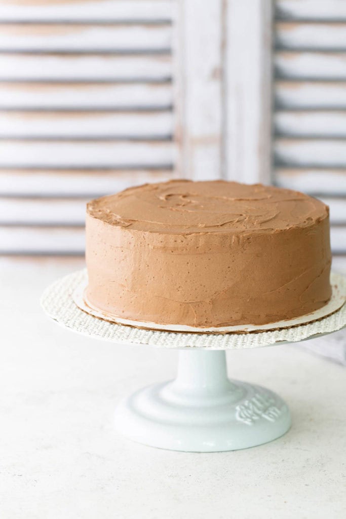 A round chocolate-frosted cake sits on a white cake stand against a light background.