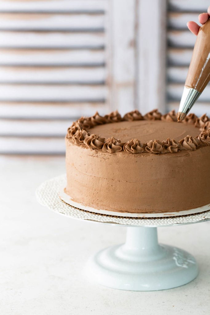 A hand decorates the top edge of a round chocolate cake with piped chocolate frosting on a white cake stand.