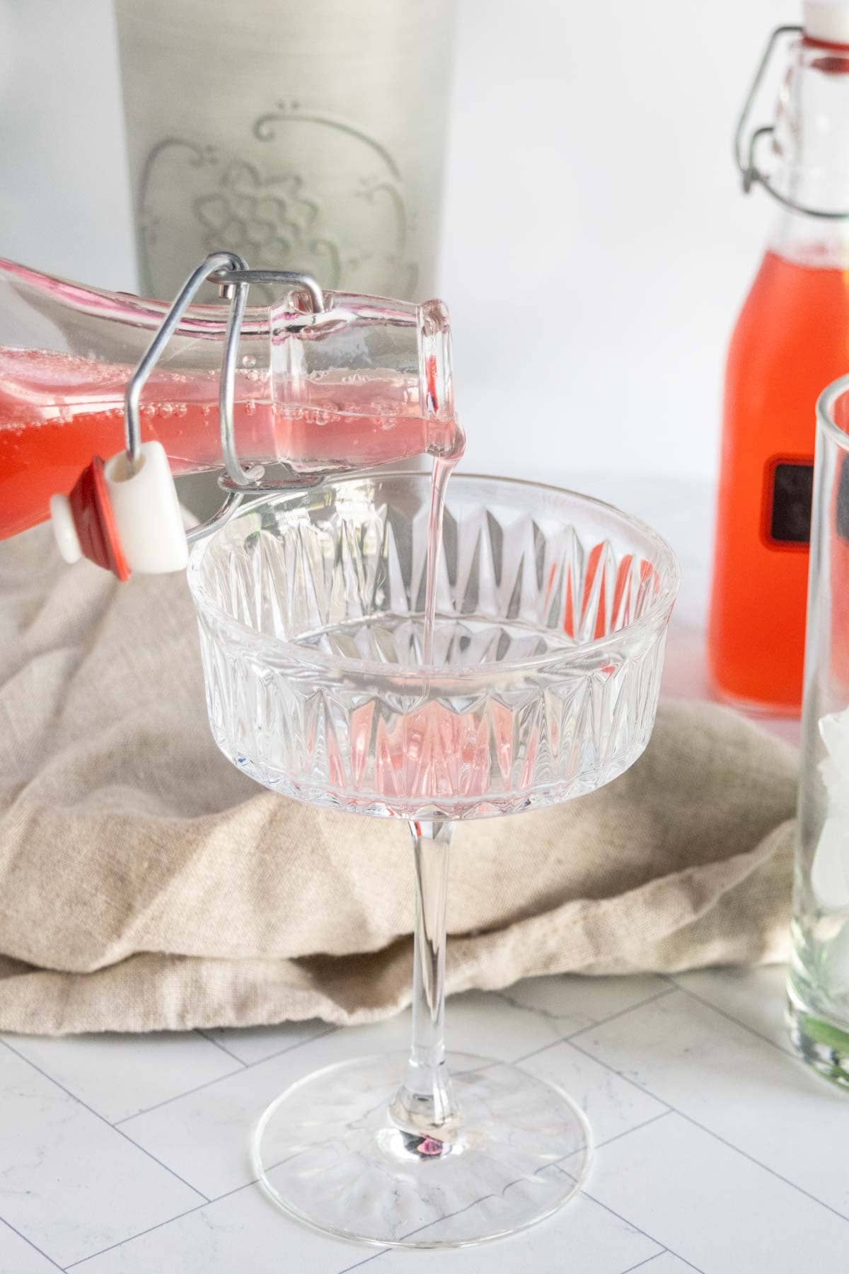 A hand pours pink liquid from a glass bottle into a stemmed crystal glass, with bottles and a beige cloth in the background.