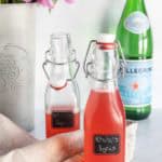 Two glass bottles labeled "Rhubarb Syrup" sit on a countertop next to a beige cloth, with a bottle of San Pellegrino and pink flowers in the background.