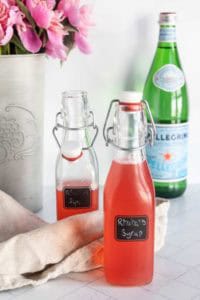 Two glass bottles labeled "Rhubarb Syrup" sit on a countertop next to a beige cloth, with a bottle of San Pellegrino and pink flowers in the background.