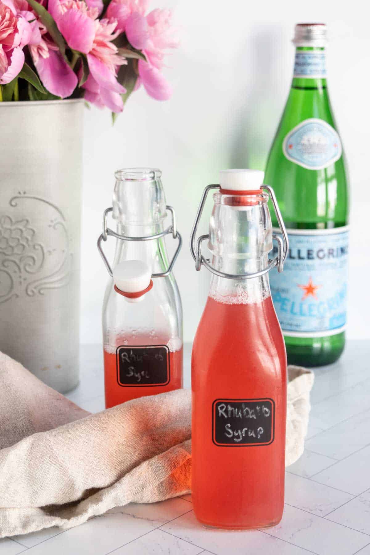 Two glass bottles labeled "Rhubarb Syrup" sit on a countertop next to a beige cloth, with a bottle of San Pellegrino and pink flowers in the background.