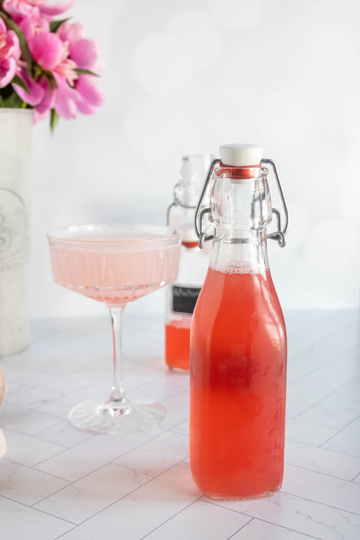 A glass bottle filled with pink liquid stands in front of a matching pink drink in a cocktail glass, with flowers and another bottle in the background.