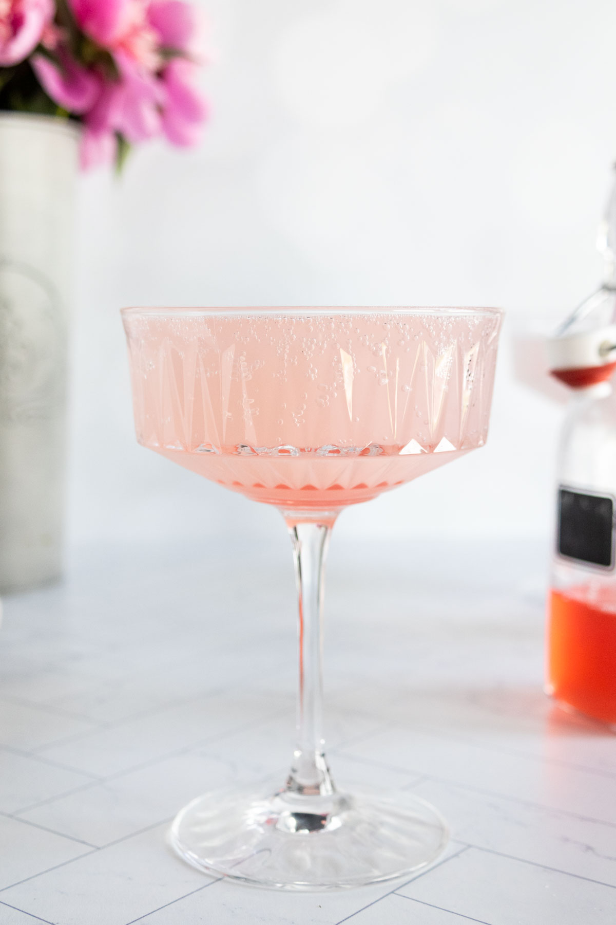 A coupe glass filled with a pale pink cocktail sits on a white surface, with blurred flowers and a small bottle in the background.