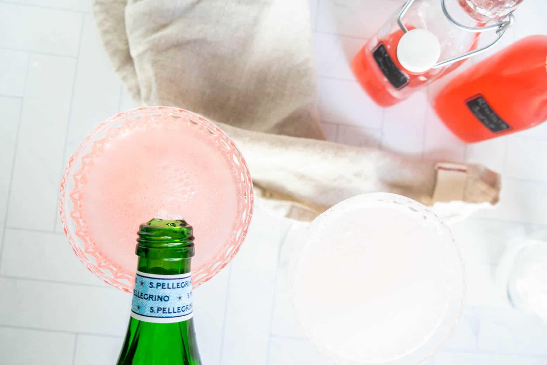 A bottle of San Pellegrino being poured into a pink cocktail glass, with another filled glass and bottles of pink liquid on a white surface in the background.