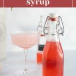 A glass bottle of pink rhubarb syrup stands next to a filled cocktail glass and another bottle in the background. The label reads “rhubarb syrup.”.
