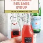 Two glass bottles filled with pink rhubarb syrup sit on a table, with a bottle of sparkling water and a beige cloth nearby. A recipe sign is displayed above the bottles.