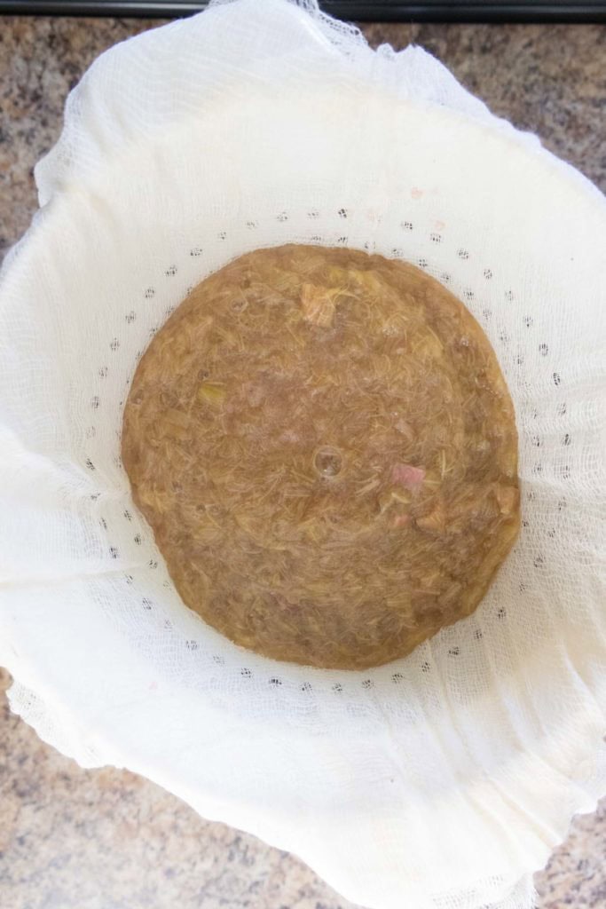 A round mass of cooked, strained rhubarb pulp sits on a cheesecloth-lined strainer over a countertop.