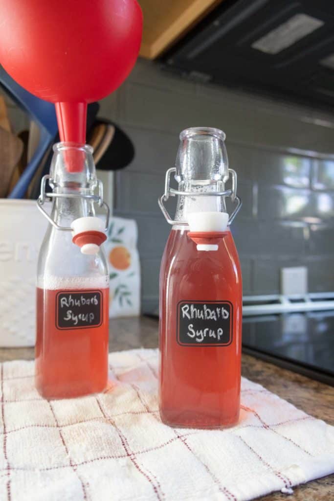 Two glass bottles labeled "Rhubarb Syrup" sit on a kitchen towel; one bottle has a red funnel inserted in the top.