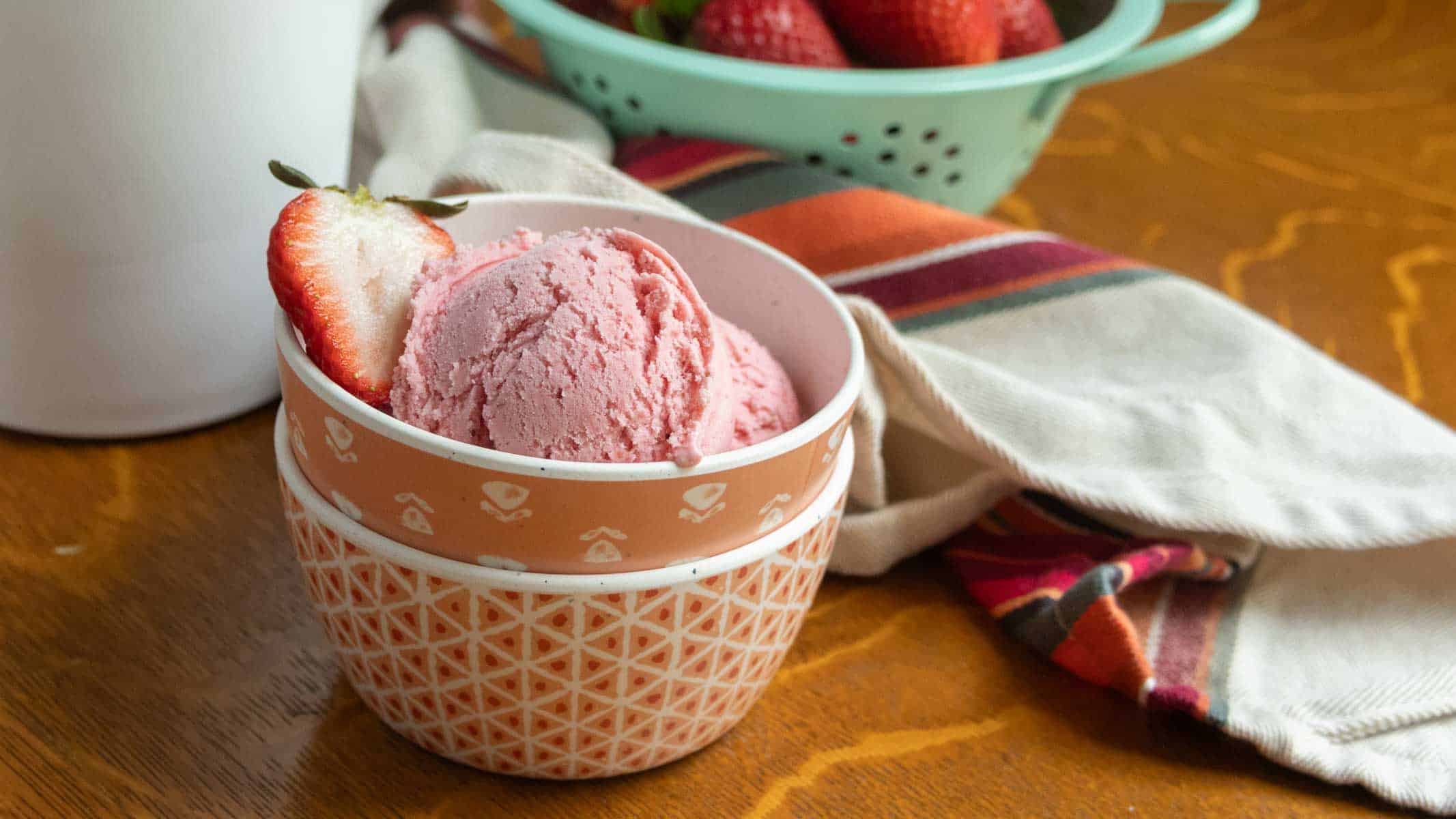 A bowl of strawberry ice cream garnished with a sliced strawberry, with a colander of strawberries and a striped cloth in the background on a wooden table.