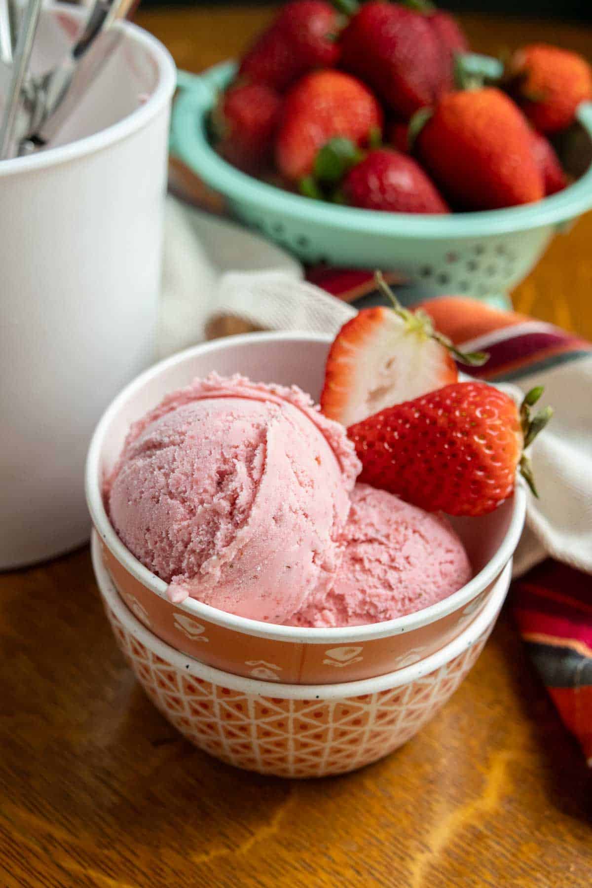 A bowl of strawberry ice cream with two fresh strawberries on the side, a white container with spoons, and a bowl of strawberries in the background.