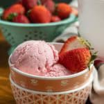 A bowl of strawberry ice cream with fresh strawberries on top, placed on a wooden surface with a bowl of whole strawberries in the background.