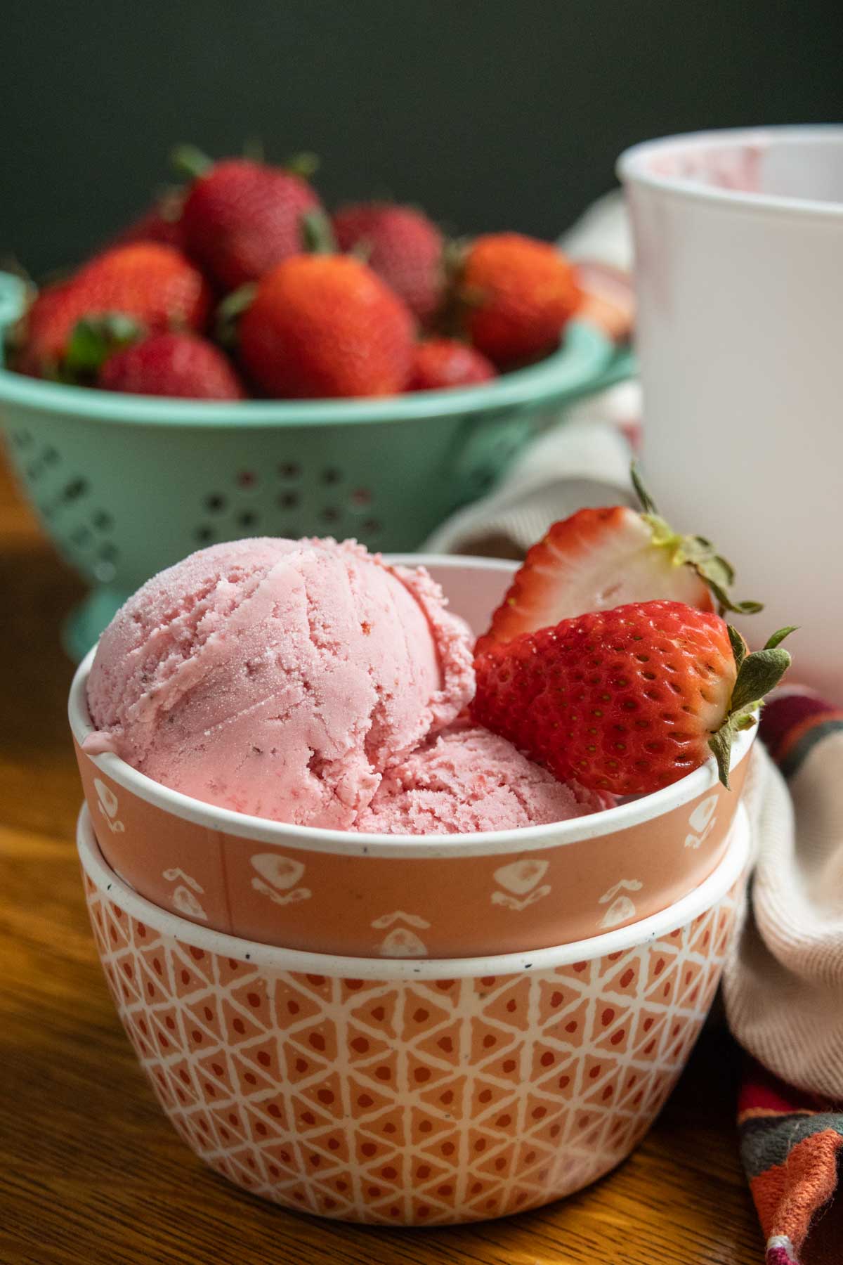 A bowl of strawberry ice cream with fresh strawberries on top, placed on a wooden surface with a bowl of whole strawberries in the background.