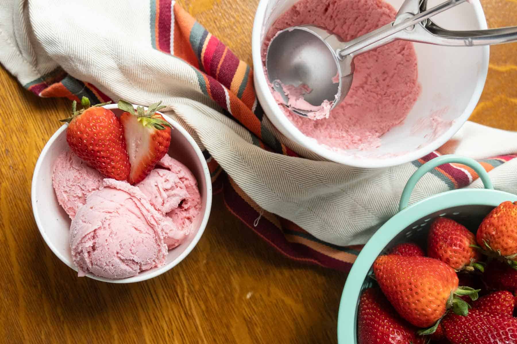 A bowl of strawberry ice cream with fresh strawberries, a metal ice cream scoop in a white bowl, and a container of strawberries on a wooden table.
