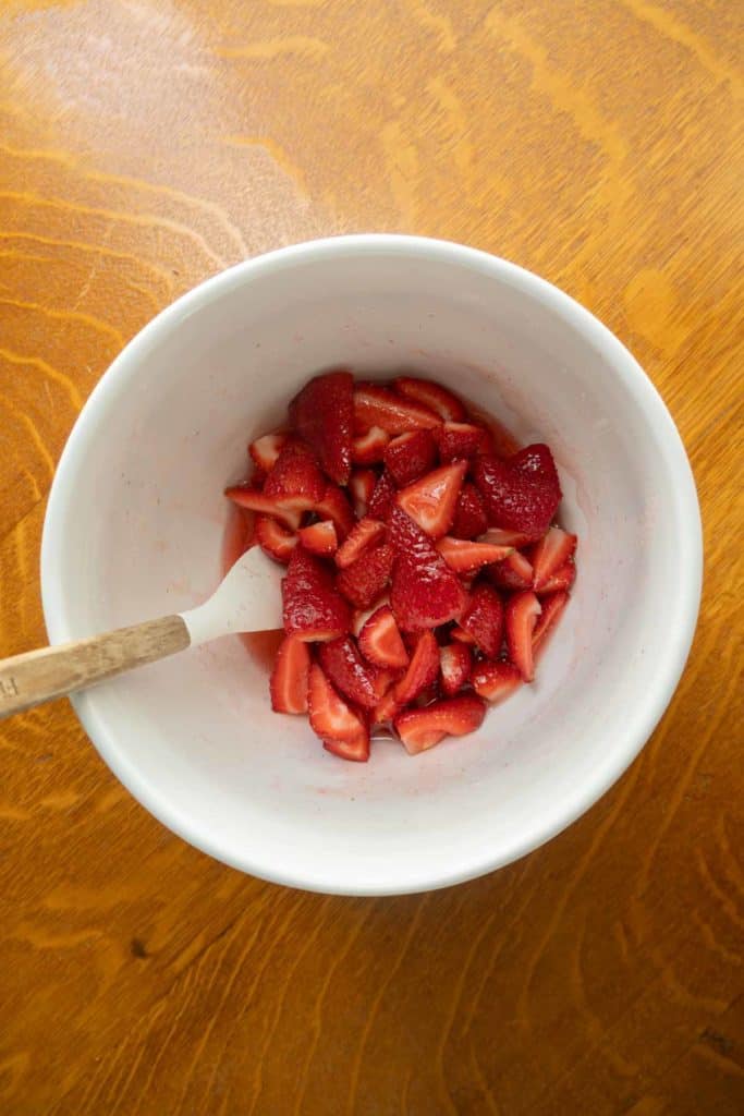 A white bowl containing sliced strawberries with a white spatula on a wooden surface.
