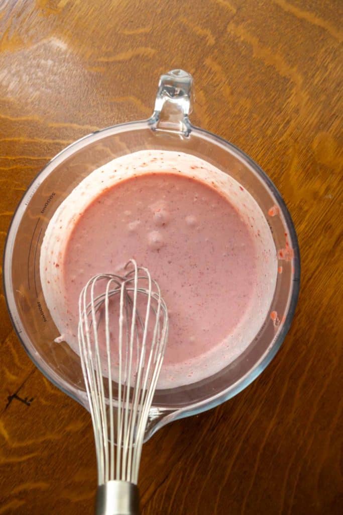 A metal whisk in a glass bowl containing a pink batter or mixture sits on a wooden surface.