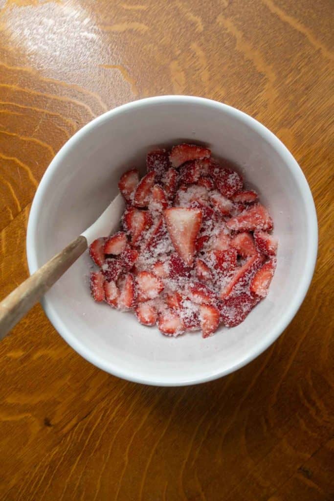 A white bowl containing sliced strawberries coated in sugar, with a wooden spoon resting inside, sits on a wooden surface.