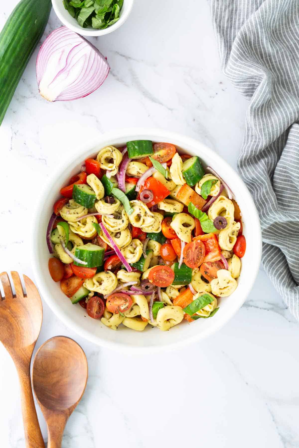 A white bowl filled with tortellini pasta salad, cherry tomatoes, cucumbers, red onions, olives, and herbs, on a marble surface with wooden utensils and a striped cloth nearby.