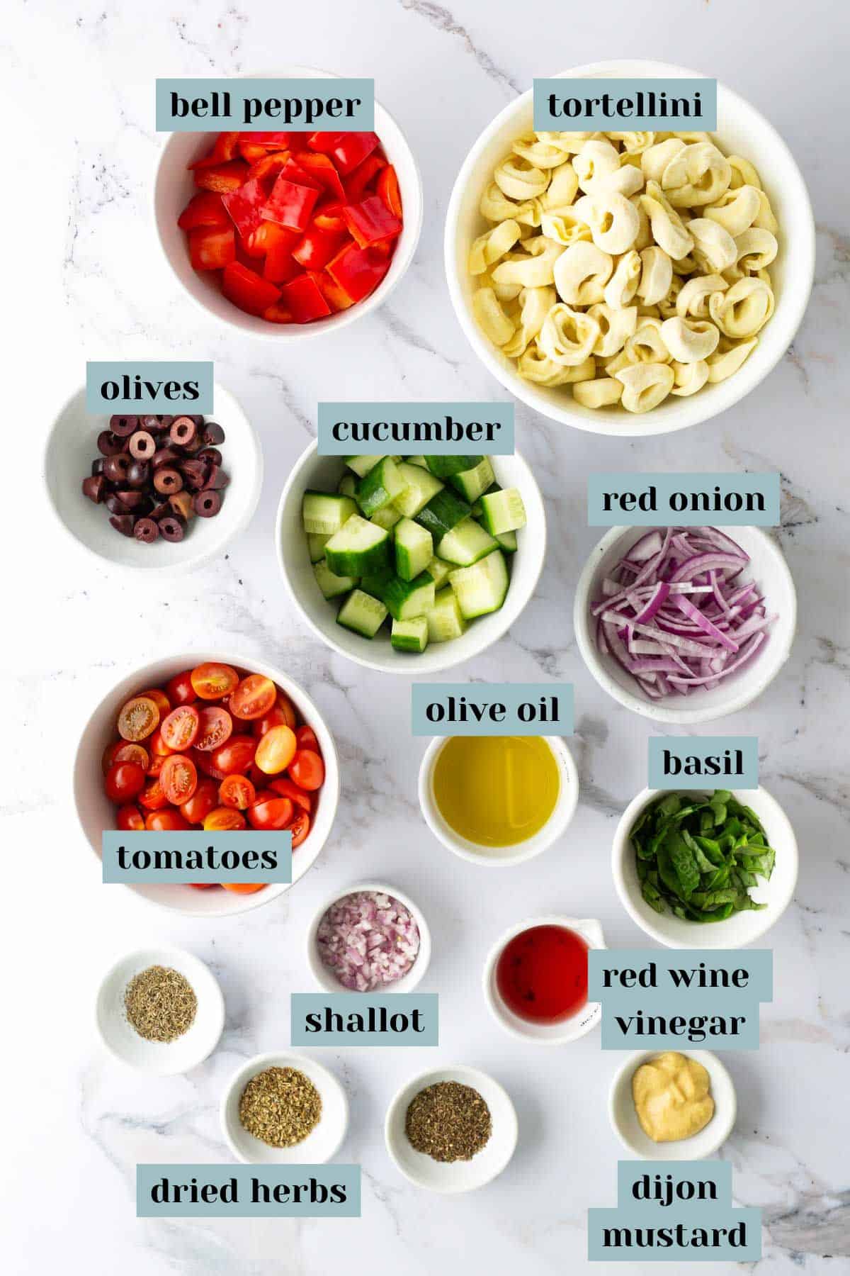 Bowls of chopped bell pepper, tortellini, olives, cucumber, red onion, tomatoes, olive oil, basil, red wine vinegar, shallot, dried herbs, and Dijon mustard arranged on a marble surface.