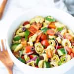 A white bowl filled with vegetable tortellini pasta salad, featuring cherry tomatoes, cucumbers, red onion, bell peppers, olives, and herbs. Wooden utensils are nearby.