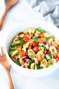 A white bowl filled with vegetable tortellini pasta salad, featuring cherry tomatoes, cucumbers, red onion, bell peppers, olives, and herbs. Wooden utensils are nearby.