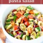 A white bowl filled with tortellini pasta salad, featuring cherry tomatoes, cucumbers, red onions, olives, and bell peppers.