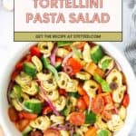 A white bowl filled with tortellini pasta salad, featuring cherry tomatoes, cucumber, red onion, olives, and bell peppers, viewed from above.