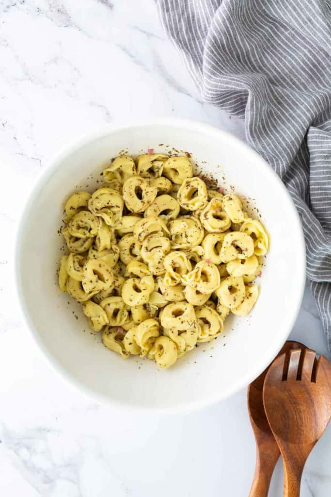 A white bowl filled with seasoned tortellini pasta sits on a marble surface next to a striped gray cloth and wooden salad utensils.