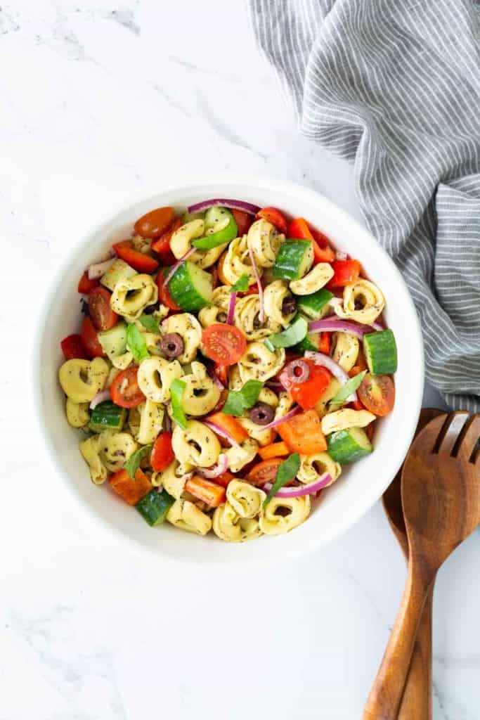 A white bowl filled with tortellini pasta salad with tomatoes, cucumbers, red onions, bell peppers, and olives sits on a marble surface next to a striped cloth and wooden salad servers.