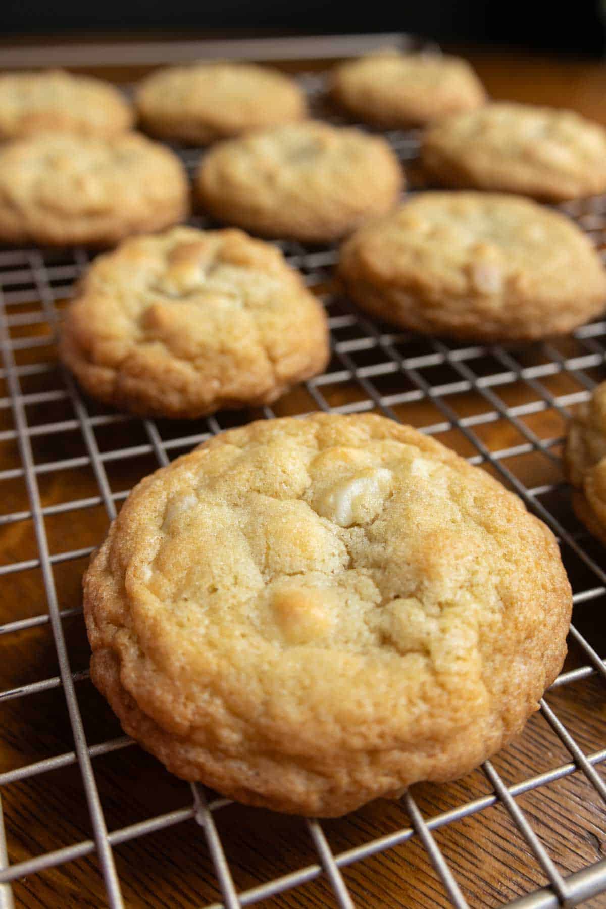 Close-up of several freshly baked cookies with white chocolate chips cooling on a wire rack over a wooden surface.
