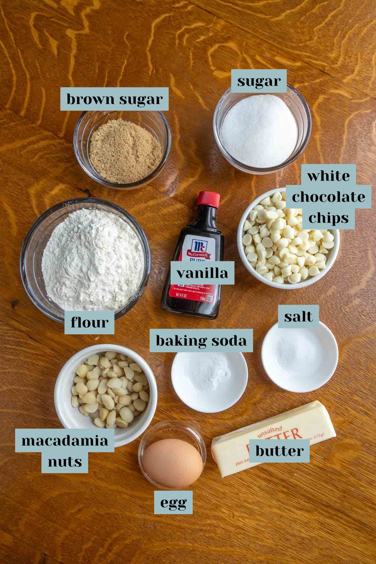 Ingredients for baking are arranged on a wooden surface, including flour, brown sugar, white sugar, white chocolate chips, macadamia nuts, an egg, butter, vanilla, baking soda, and salt.
