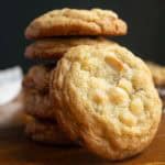 A stack of cookies with white chocolate chips on a wooden surface, with a few white chocolate chips in the foreground.