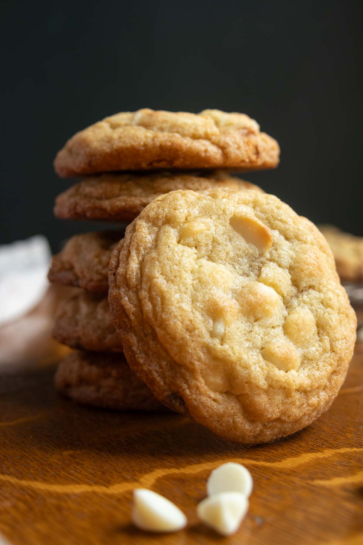 A stack of cookies with white chocolate chips on a wooden surface, with a few white chocolate chips in the foreground.
