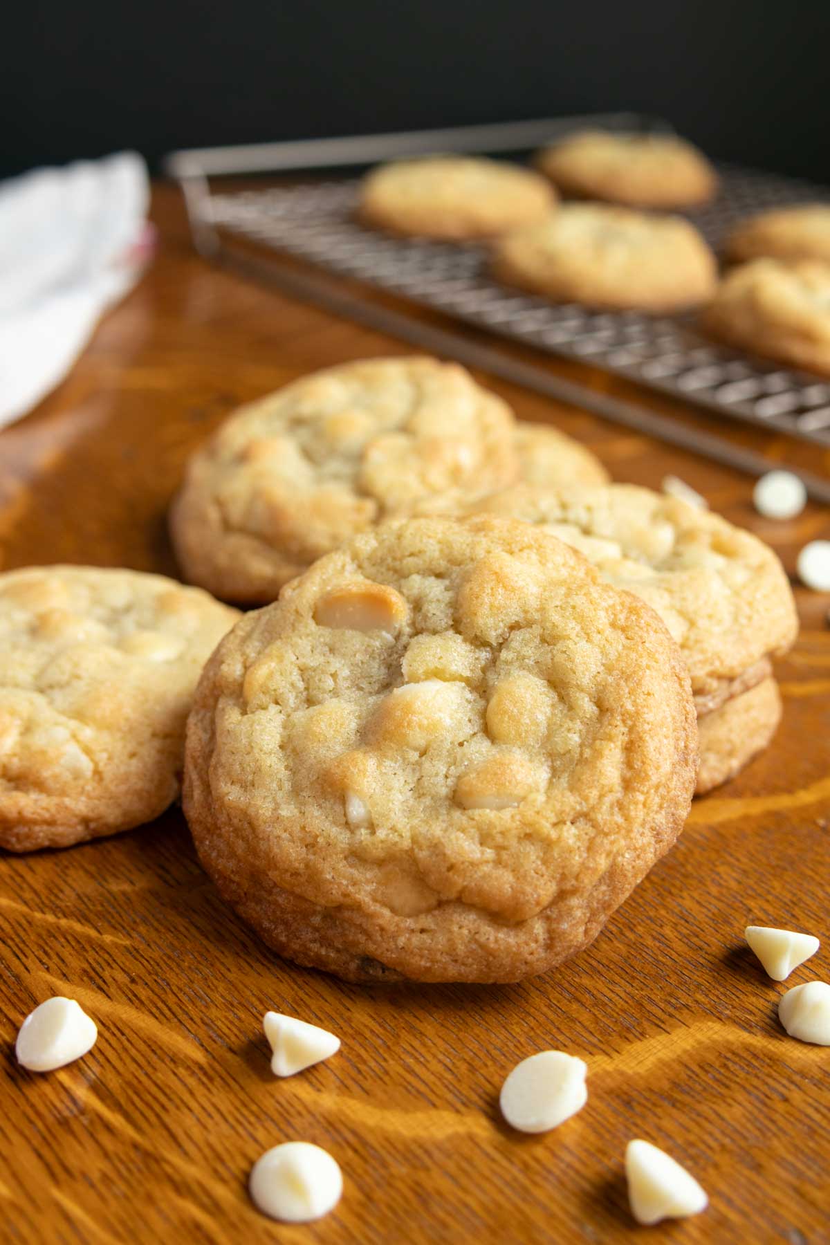 A close-up of white chocolate chip cookies on a wooden surface, with more cookies cooling on a wire rack in the background. White chocolate chips are scattered nearby.
