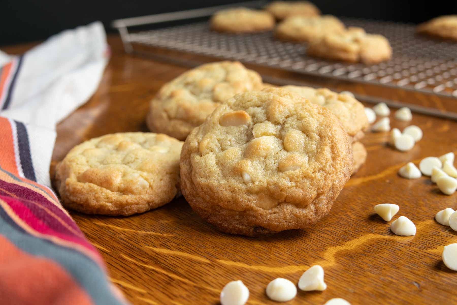 A close-up of white chocolate chip cookies on a wooden surface with scattered white chocolate chips, a striped cloth, and a cooling rack in the background.