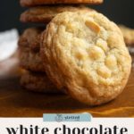 A stack of white chocolate macadamia nut cookies on a wooden surface, with one cookie leaning against the stack.
