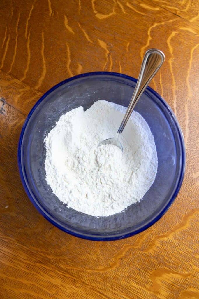 A blue glass bowl filled with white flour and a metal spoon, placed on a wooden surface.