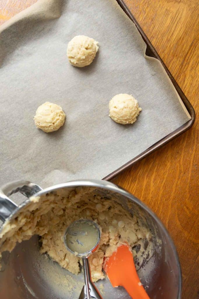 Three scoops of cookie dough on a parchment-lined baking sheet with a mixing bowl, dough, scoop, and orange spatula on a wooden surface.