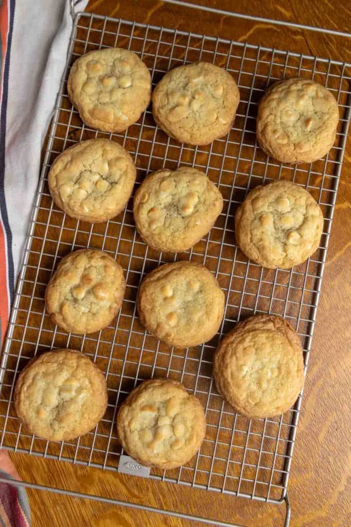 Eleven baked cookies with white chocolate chips cooling on a metal wire rack, placed on a wooden surface next to a striped cloth.