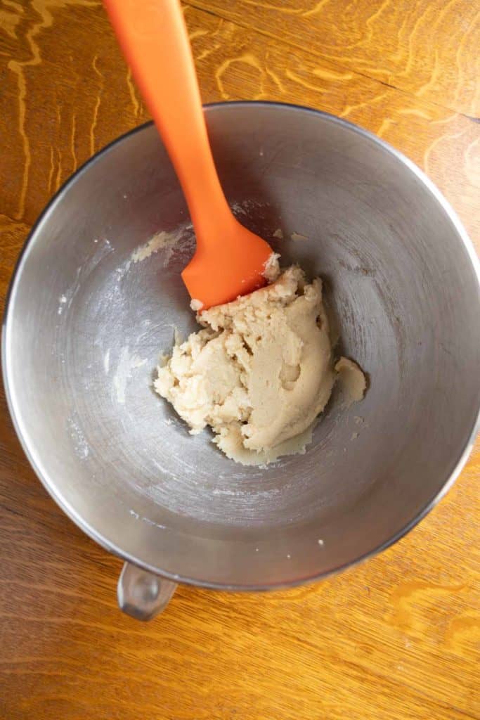 A metal mixing bowl with a small amount of dough and an orange spatula on a wooden surface.
